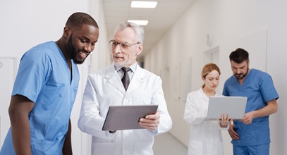 two healthcare workers discussing something in the forefront while 2 other healthcare workers look at a laptop in the background of the hallway