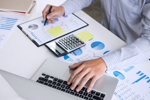 person in long sleeves typing on a laptop while review charts on a desk