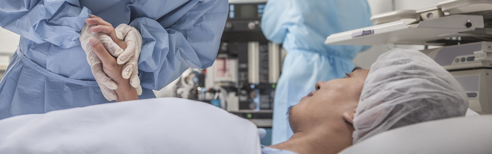 female healthcare worker in blue surgery scrubs holding the hand of a male patient laying down on a table