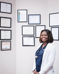 Chelsea Asanta in a white nursing coat and stethoscope standing with diplomas and accolades on the wall