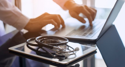 stethoscope and a tablet next to the healthcare provider typing on laptop
