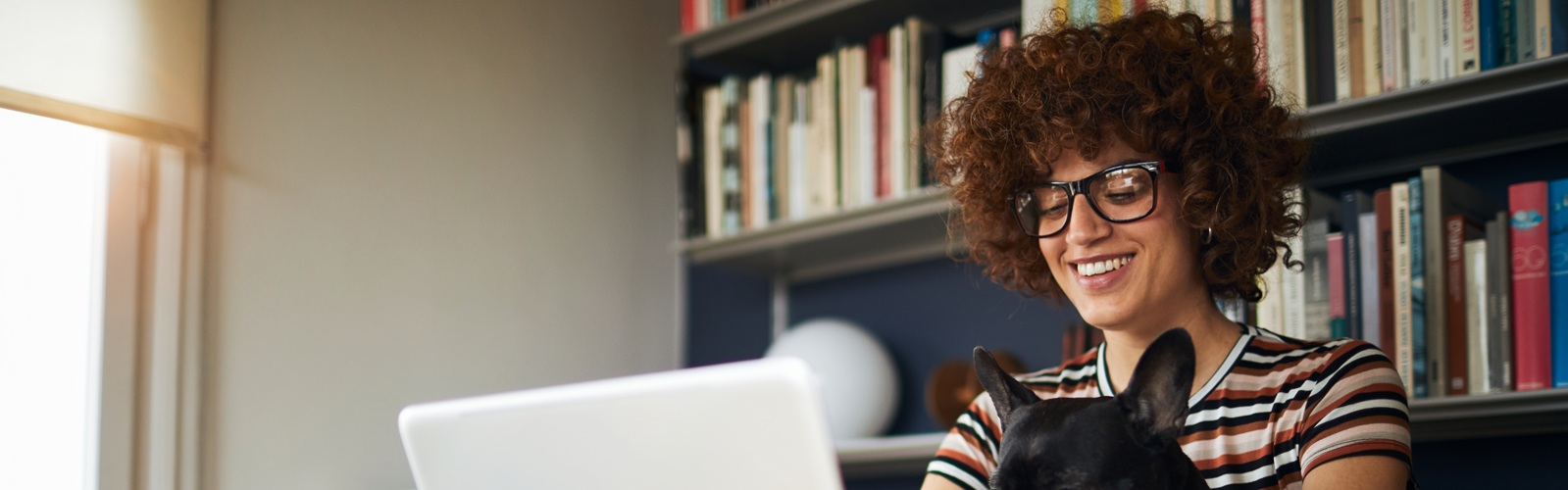 woman with brown curly hair and glasses smiling sitting at her laptop with a french bulldog on her lap