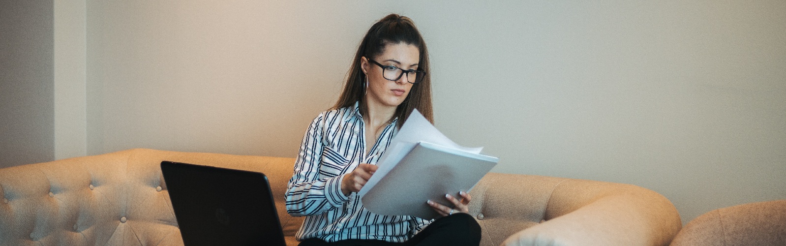 brown haired woman sitting crossed legged on a couch in a blouse and slacks with a laptop on her lap looking at papers