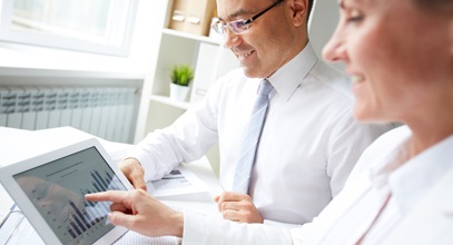 a man and a woman both in white shirts sitting together at a desk looking at a tablet screen together