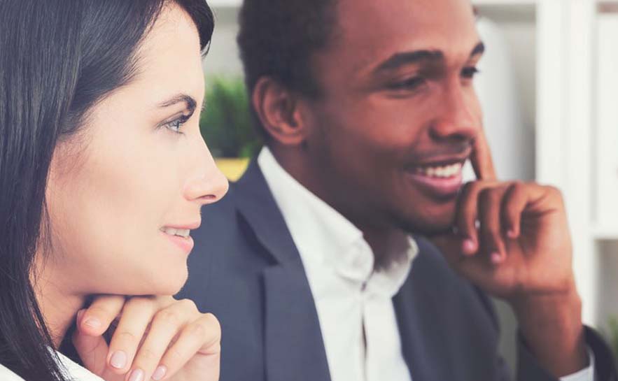 Two individuals sitting side by side. The woman on the left is wearing a white shirt with a hand on her chin, while the man on the right is in a suit with a hand on his chin.