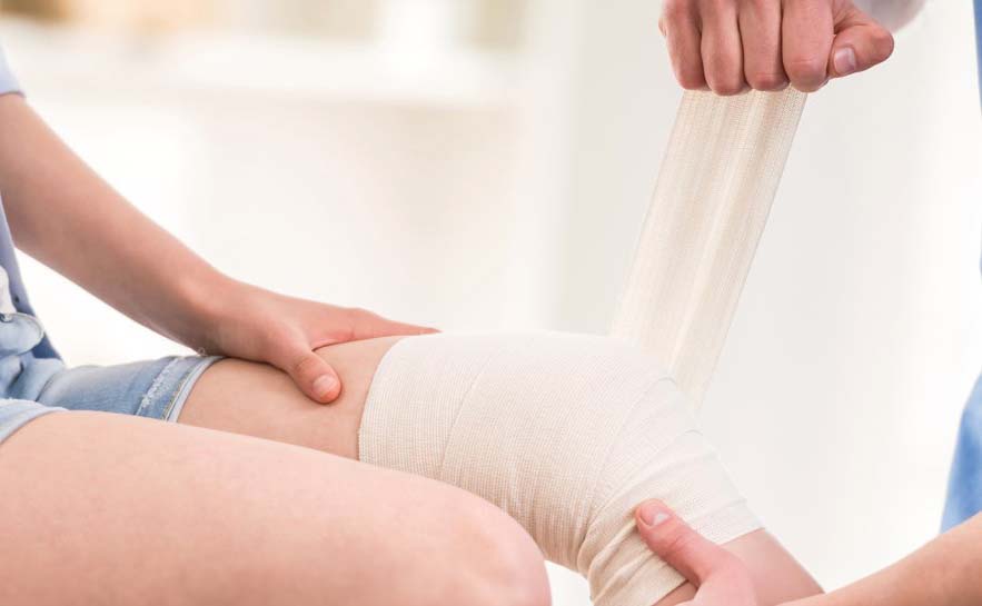 Person sitting while another wraps a white elastic bandage around their knee.
