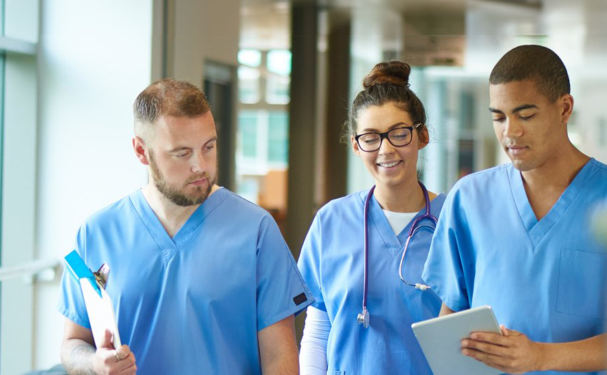 Three healthcare professionals in blue scrubs standing together in a hospital hallway. One is holding a clipboard, another has a stethoscope around the neck, and the third is holding a tablet.