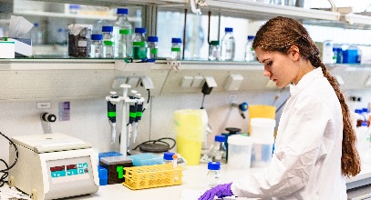 Young lady in a white lab coat with long brown hair studying a book in a pharmacy lab