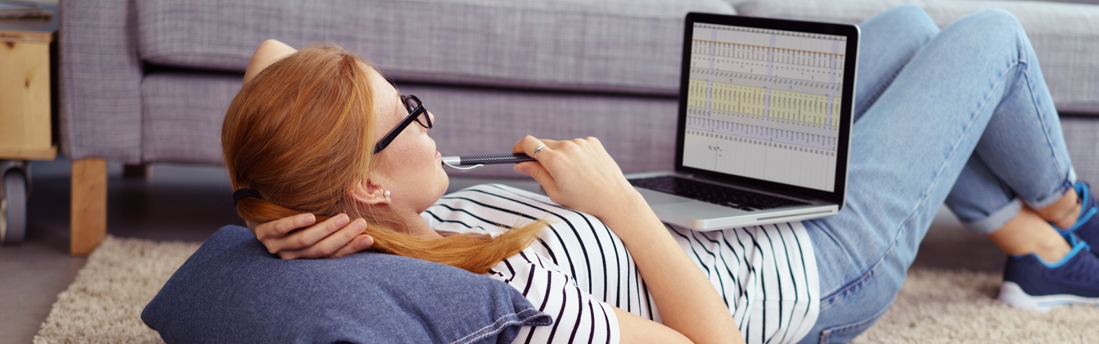 young redheaded lady laying down on the floor looking at a laptop with a notebook by her side
