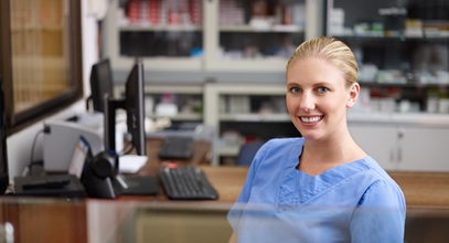 young blonde lady sitting at a desk in blue scrubs with a desktop computer