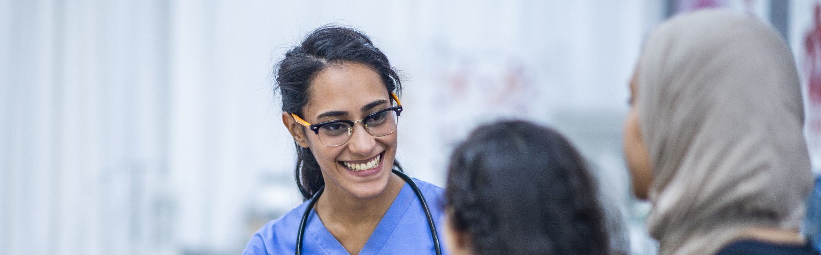 healthcare worker in blue scrubs and a stethoscope speaking to a child and her mother