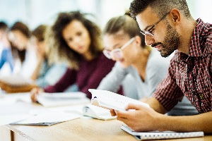 man with a slight beard and checkered button down shirt reading a book with additional classmates in the background studying